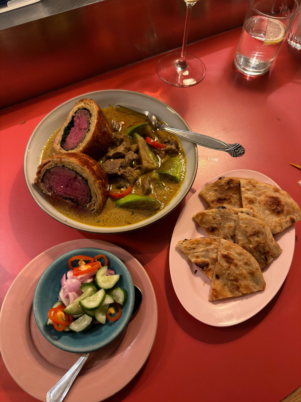 A bowl of green curry with sliced beef in pastry, next to a plate of roti and a small bowl of cucumber and chilli relish.