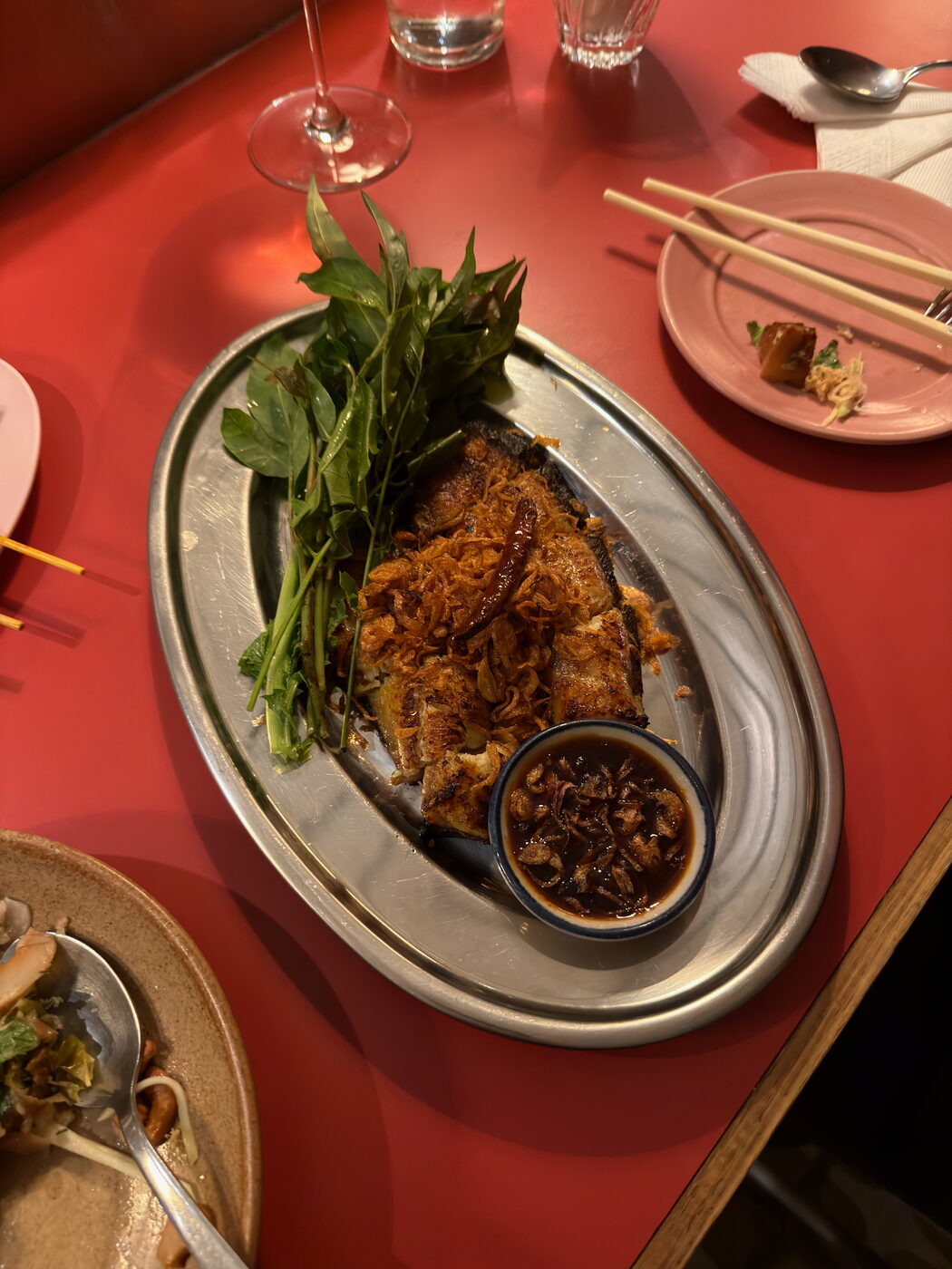 A metal platter holding a crisp-fried fish dish topped with fried shallots, served with a bunch of fresh herbs and a small bowl of dark dipping sauce.