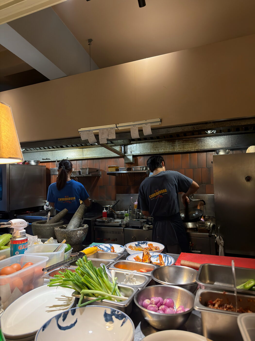 Cooks at work in Charmkrung's open kitchen, behind a counter lined with stone mortars, prepared vegetables and bowls of ingredients.